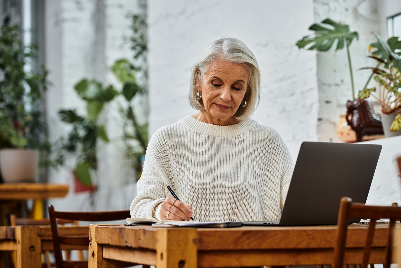 A beautiful mature woman with gray hair writes intently at a wooden table in a sunny cafe.
