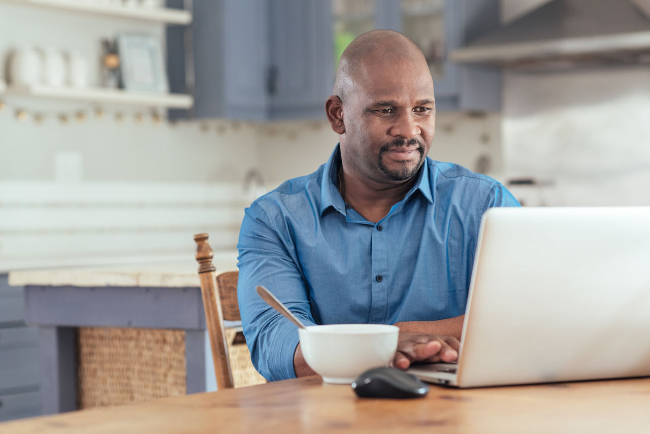 Smiling mature African man sitting at his kitchen table eating breakfast and browsing online with a laptop