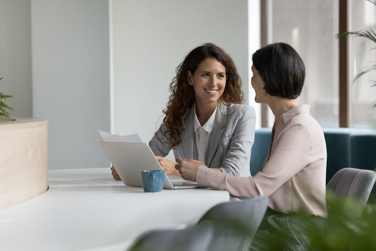 Two positive confident diverse business colleagues women talking at workplace, discussing project documents, at laptop, speaking, smiling, laughing. Mature manager consulting expert